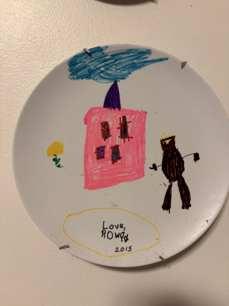 A plate with a child's drawing on it with a black figure a flower and a pink house with a blue sky.